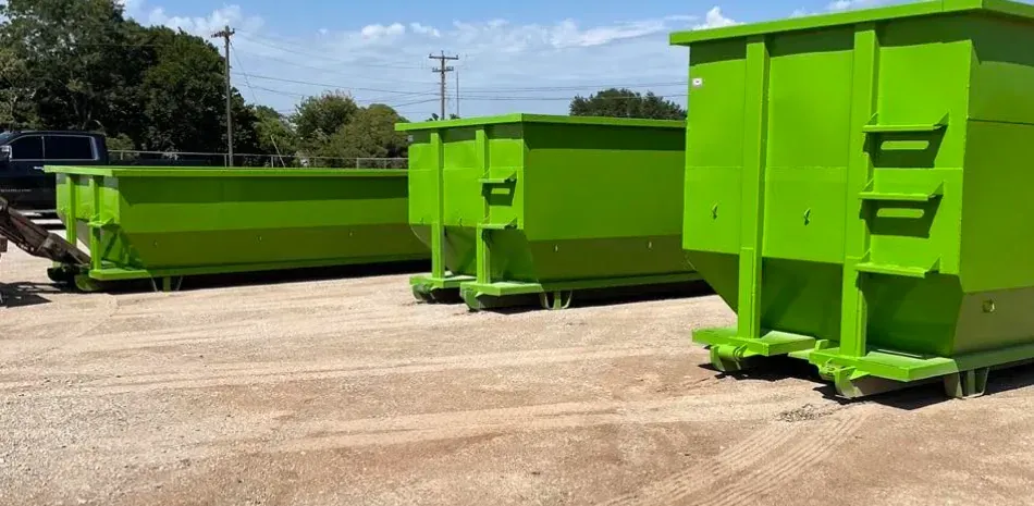 Three bright green metal dumpsters sit in a row on a gravel lot under a clear blue sky.