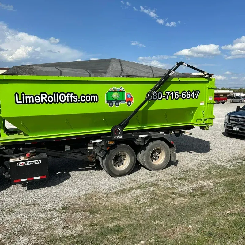 A bright green dump trailer with a covered top, parked on a gravel lot under a blue sky, displaying branding and contact.