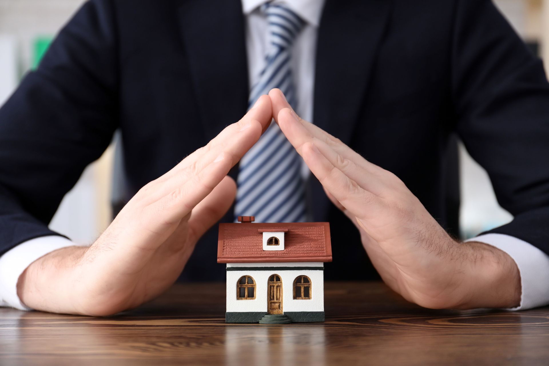 Hands forming a protective roof shape over a small model house on a wooden desk. Hands forming a protective roof shape over a small model house on a wooden desk.