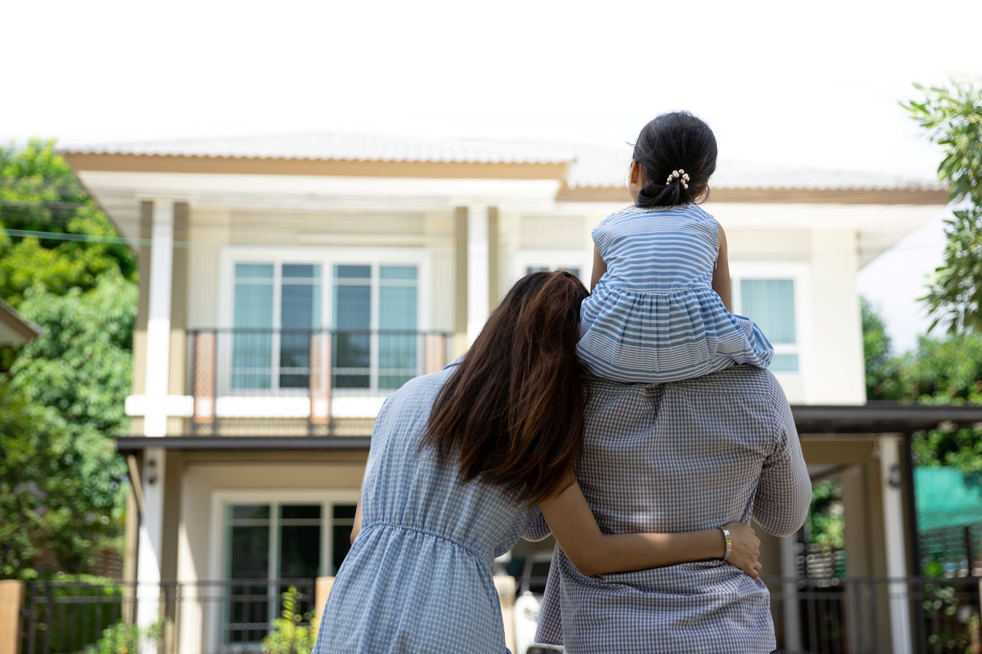 A family stands together facing their two-story home, with a child sitting on an adult’s shoulders. A family stands together facing their two-story home, with a child sitting on an adult’s shoulders.
