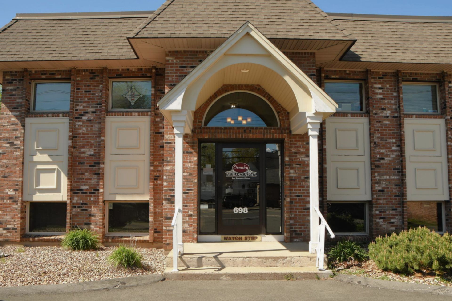Brick building with entry door, windows, and a covered porch; bushes in front.