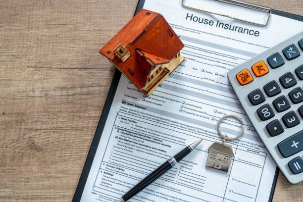 A house insurance form on a clipboard with a small model home, pen, keychain, and a calculator