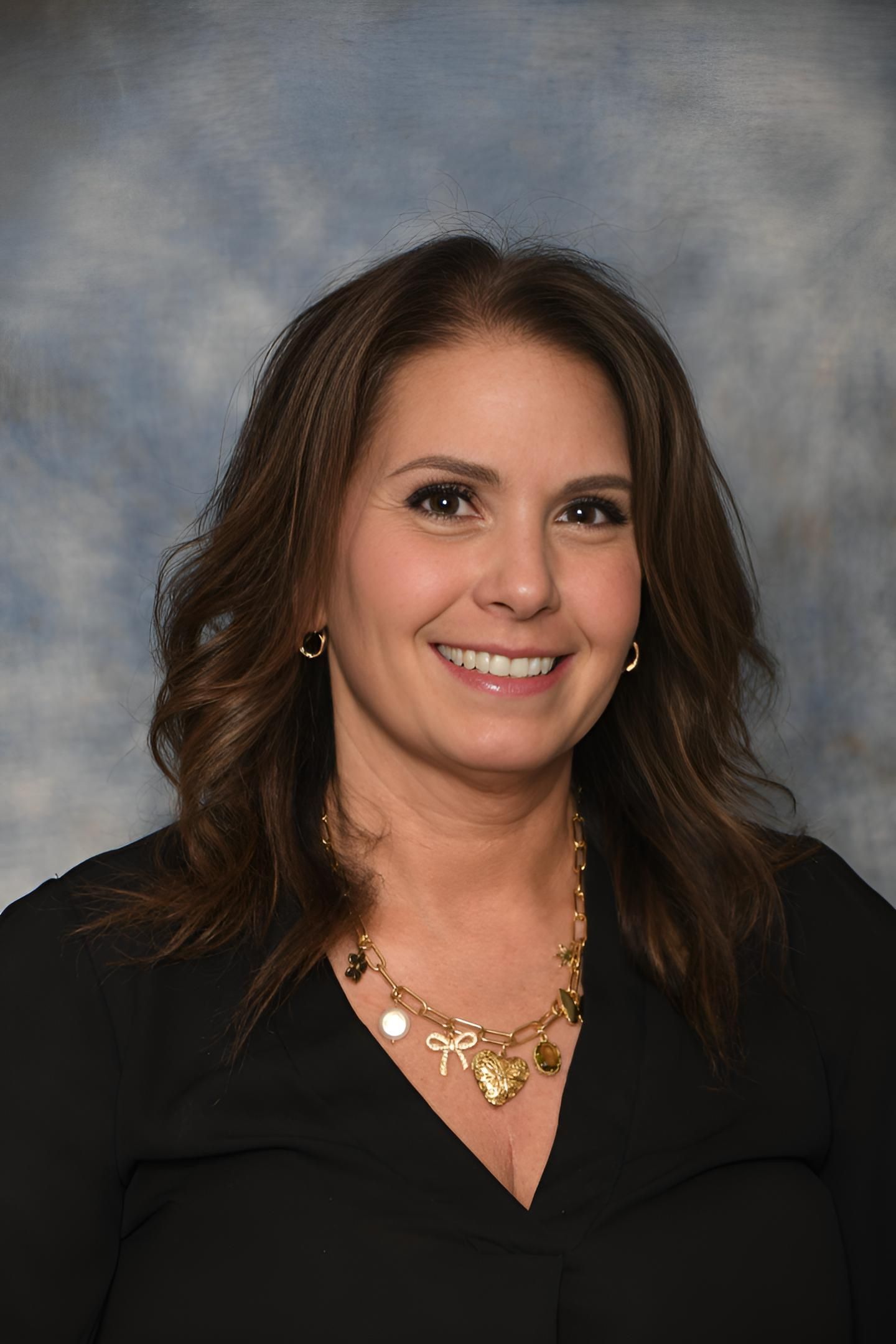 Woman with brown hair smiling, wearing a black top and gold necklace, against a blue mottled background.