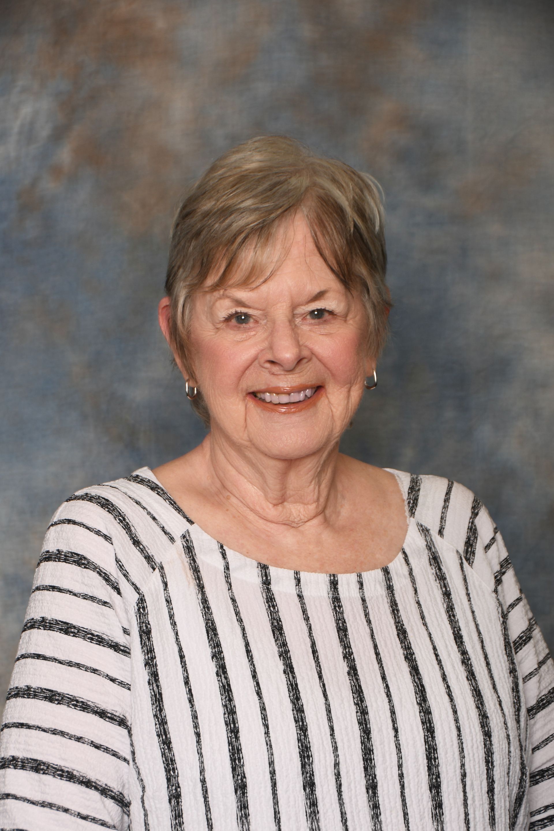 Older woman with short white hair, smiling. Wearing a black and white striped shirt against a blue and brown background.