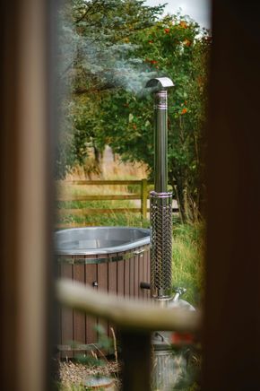 Outdoor shower beside a round tub, viewed through a window with dark frame; greenery in the background