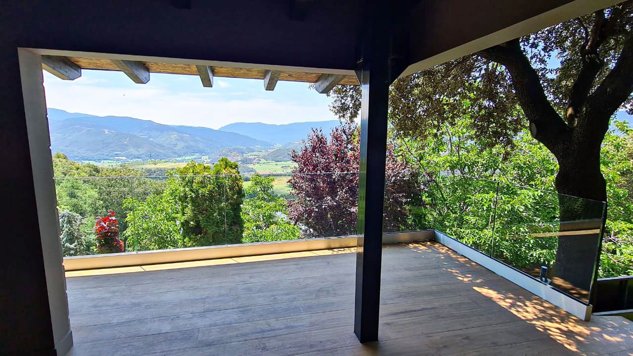 Vista des d'un porxo cobert amb vistes a una vall verda i muntanyes distants. Els arbres emmarquen la vista. Cal Serni
