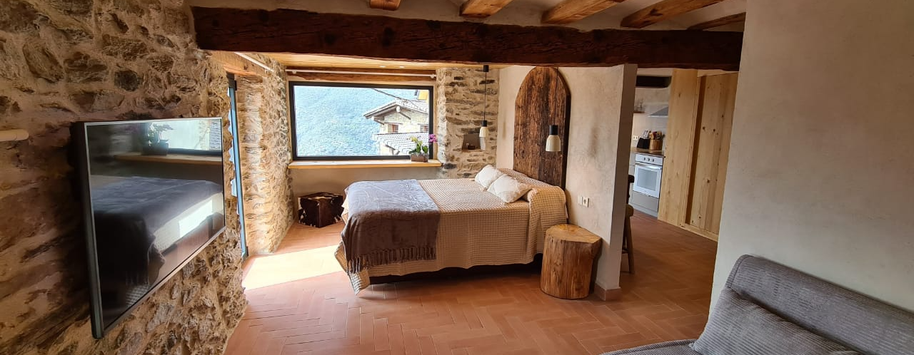 Dormitori amb paret de pedra, llit i porta oberta. La llum natural prové d'una finestra.