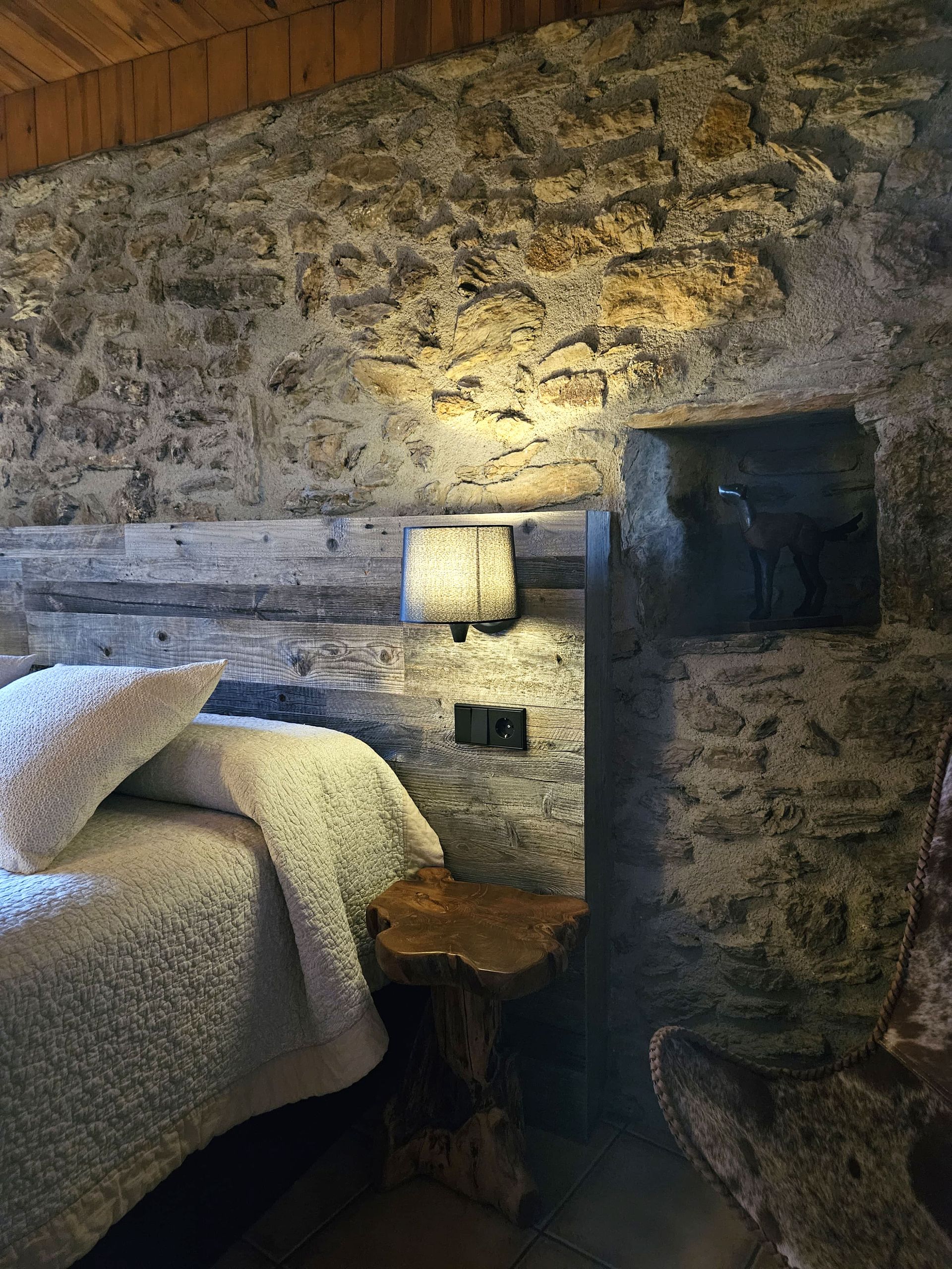 Dormitori amb parets de pedra, marc de llit de fusta, roba de llit texturitzada i tauleta auxiliar de fusta.