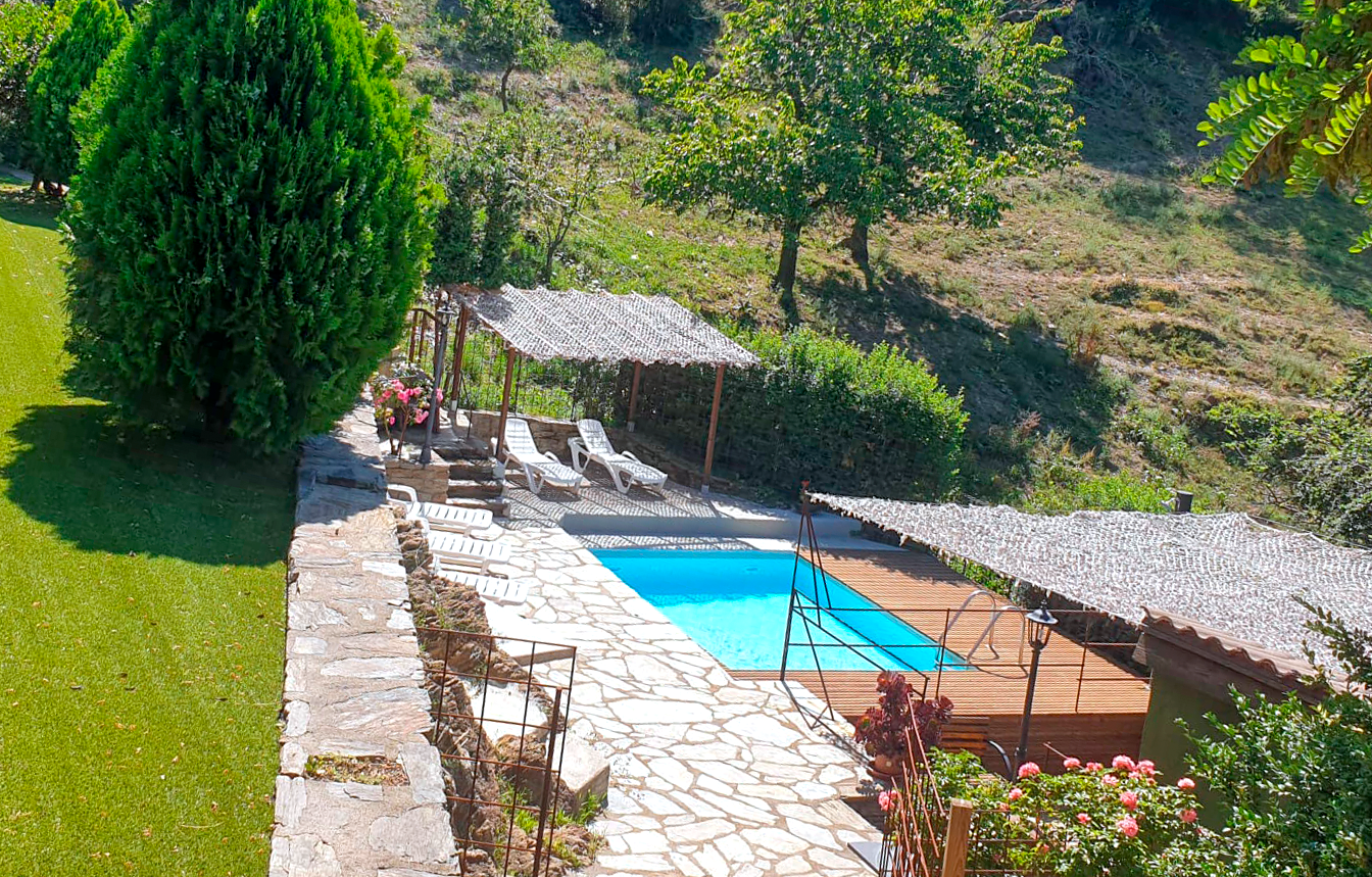 Piscina envoltada d'un pati de pedra, gespa i vegetació al vessant d'un turó, amb zones de descans cobertes. Cal Serni