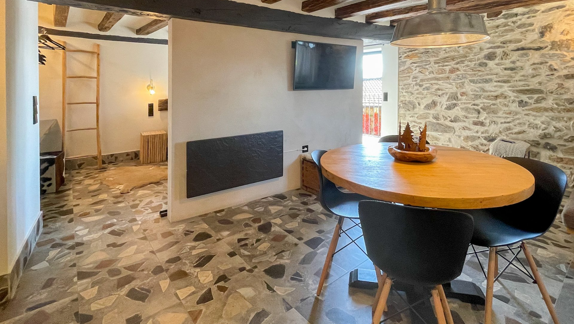 Interior acollidor amb taula de menjador, televisor, paret de pedra i dormitori visibles al fons. Cal Serni