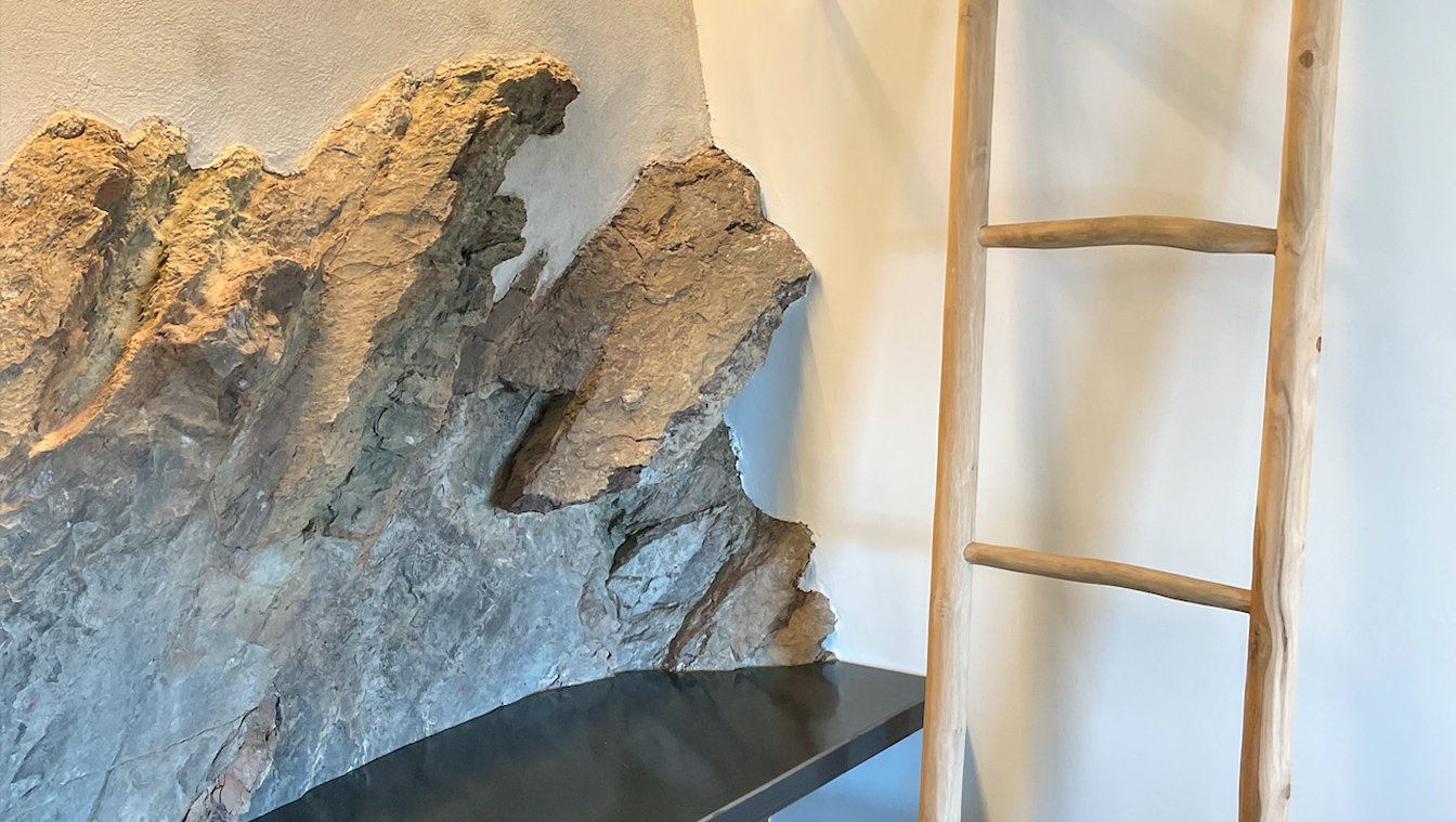 Mur de pedra rugosa al costat d'una escala de fusta contra una paret clara.