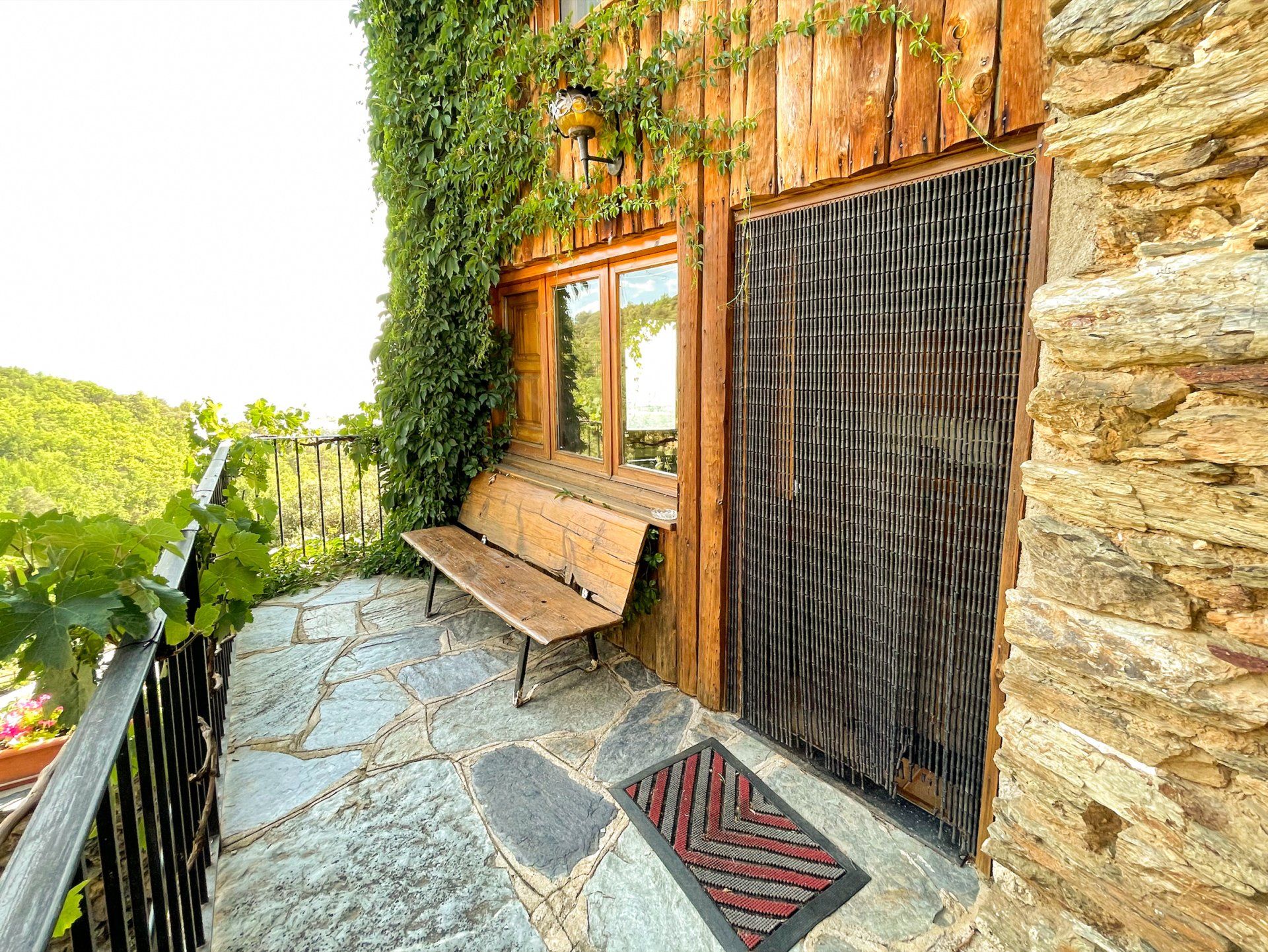 Exterior de cabana de fusta amb pati de pedra, banc i paret coberta de vinyes. Cal Serni