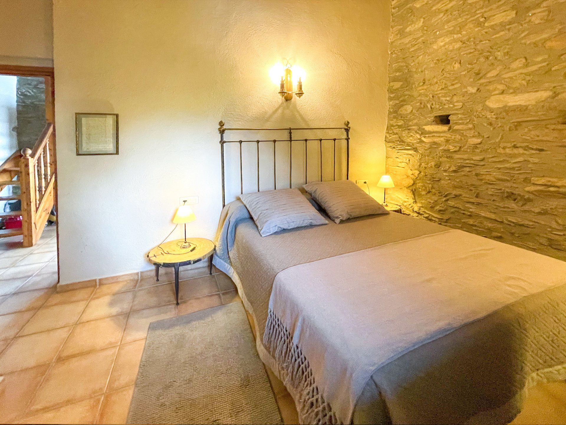 Dormitori amb llit, tauleta de nit i paret de pedra; il·luminació càlida. Cal Serni