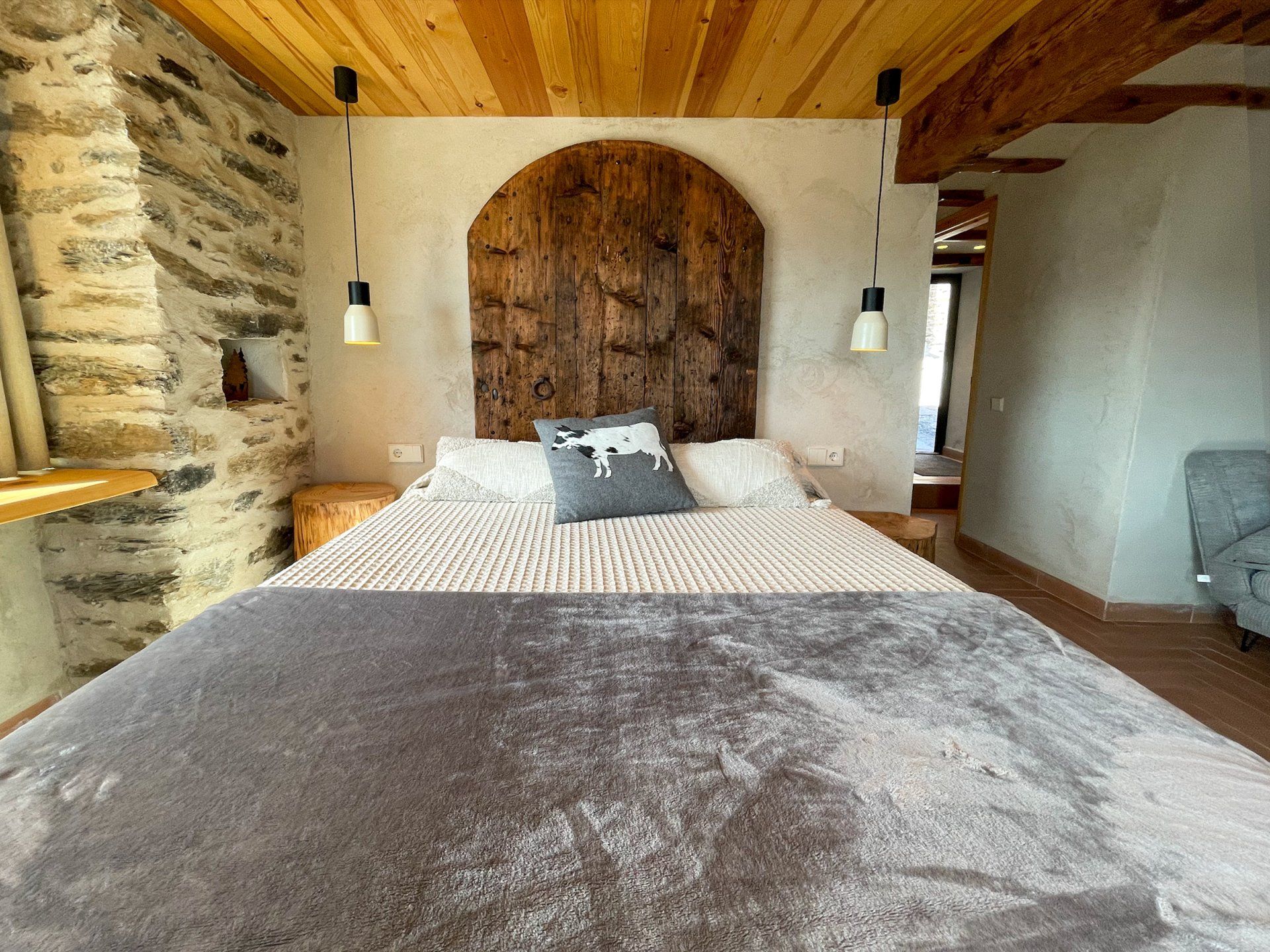 Dormitori amb capçal de fusta, paret de pedra i sostre de fusta. Llit amb manta grisa, coixí i llums penjants. Cal Serni