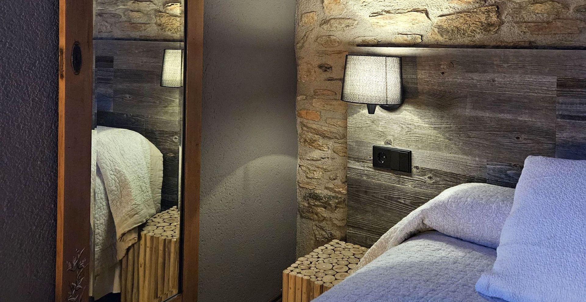 Racó del dormitori amb paret de pedra, mirall, llit i làmpada de nit. Cal Serni