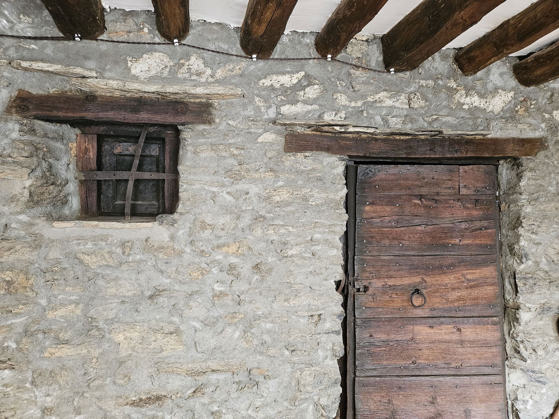Mur de pedra amb una petita finestra amb barrots i una porta de fusta; bigues rústiques al sostre. Cal Serni