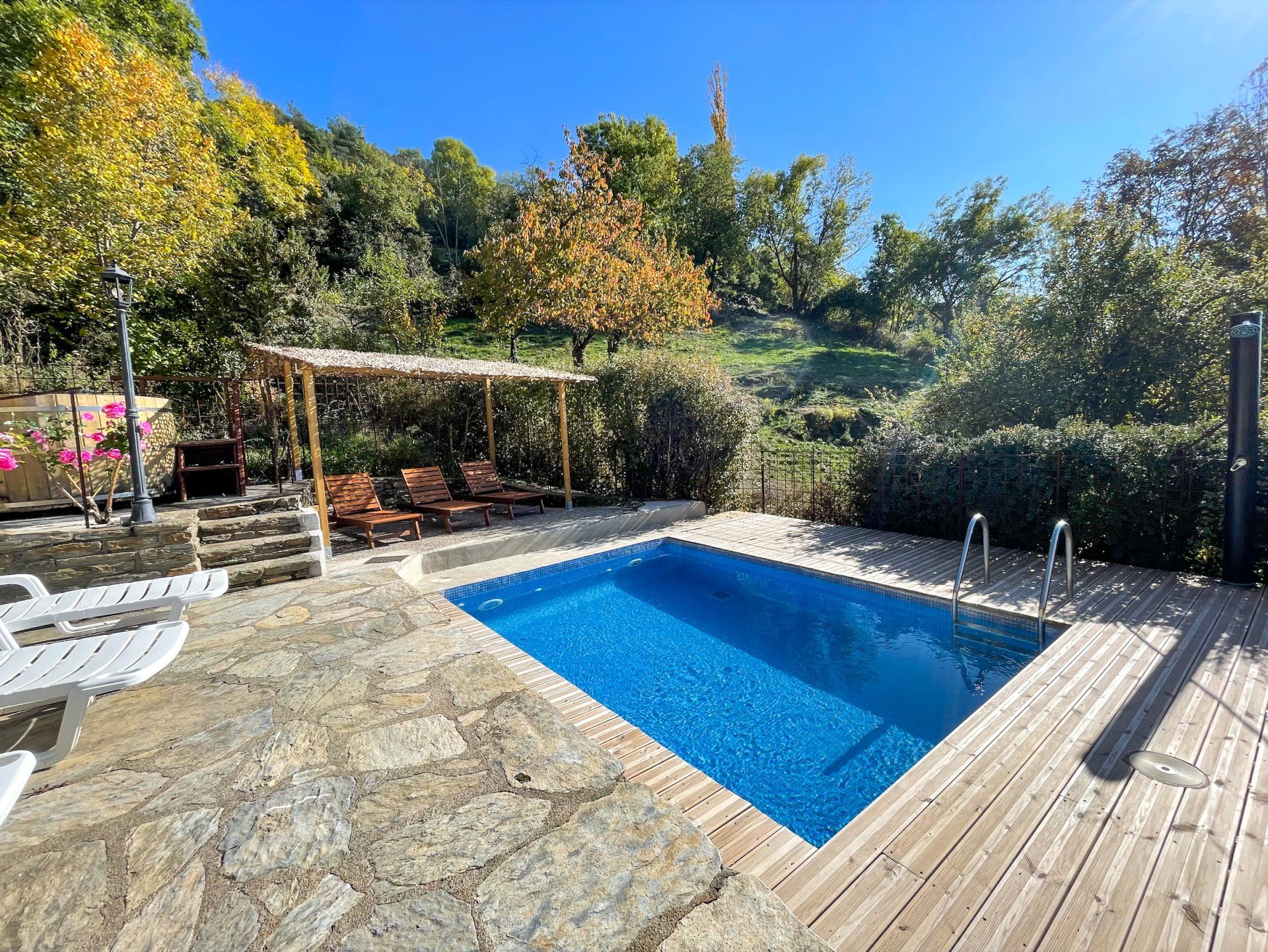 Piscina amb terrassa de fusta i gandules, envoltada de vegetació sota un cel blau. Cal Serni