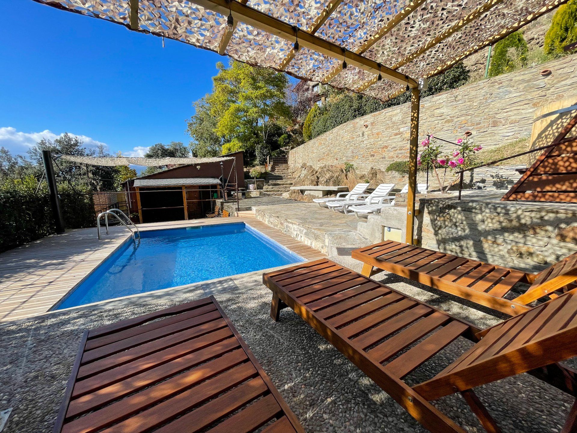 Piscina amb gandules de fusta sota una pèrgola en un pati assolellat. Cal Serni