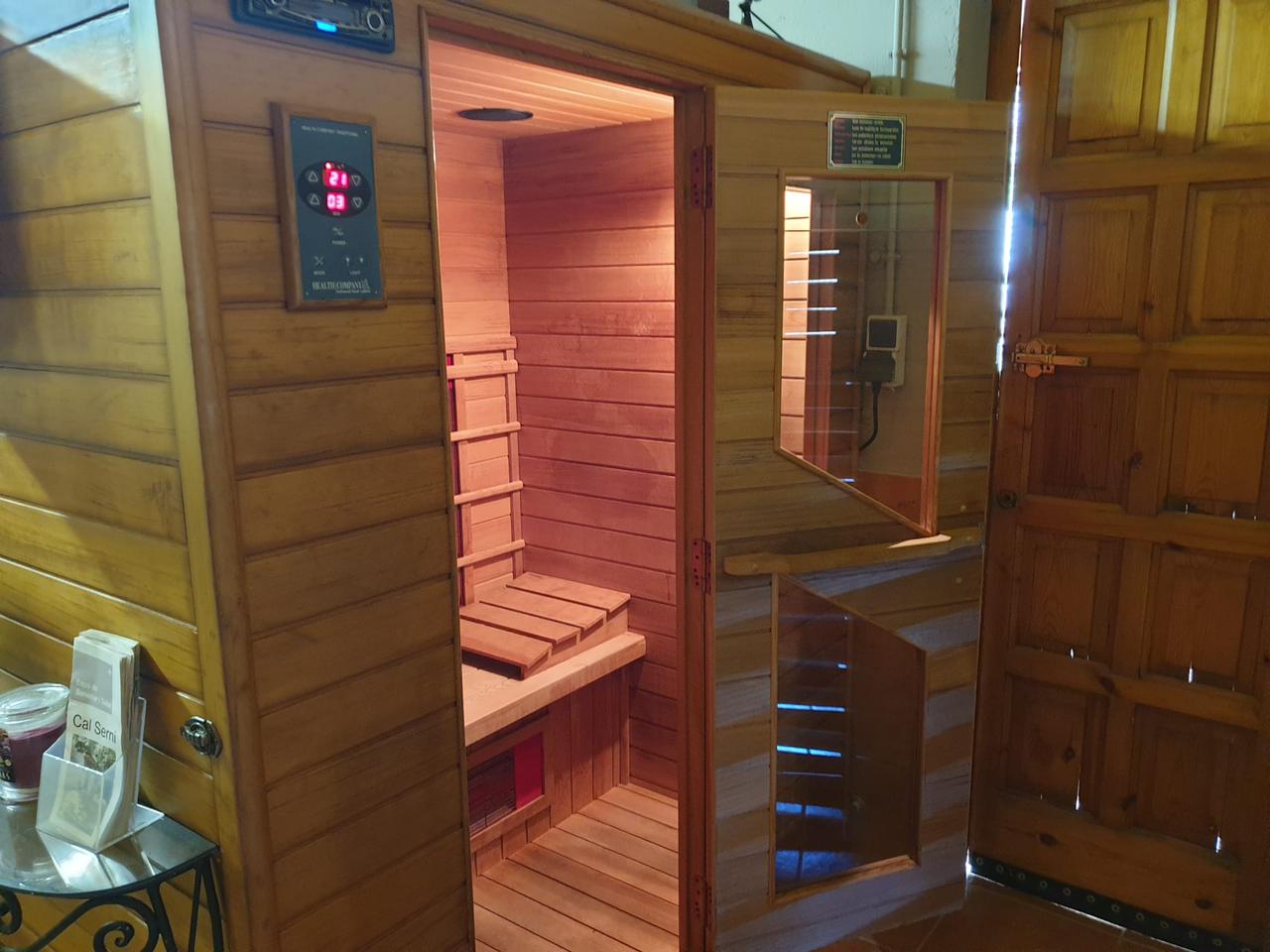 Sauna de fusta amb porta oberta, panell de control i banc a l'interior; al costat d'una porta de fusta parcialment oberta. 
