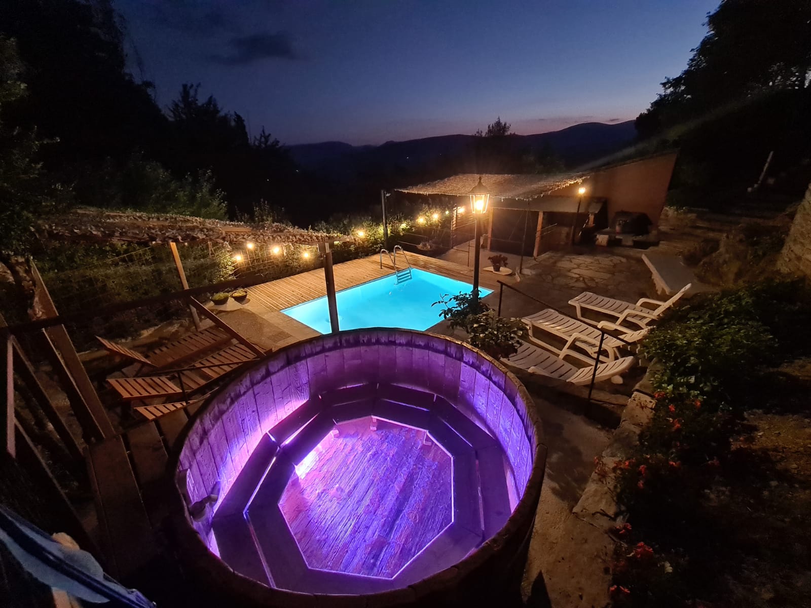 Banyera d'hidromassatge i piscina exteriors il·luminades al capvespre amb llums de corda, amb vistes a una vall. Cal Serni 