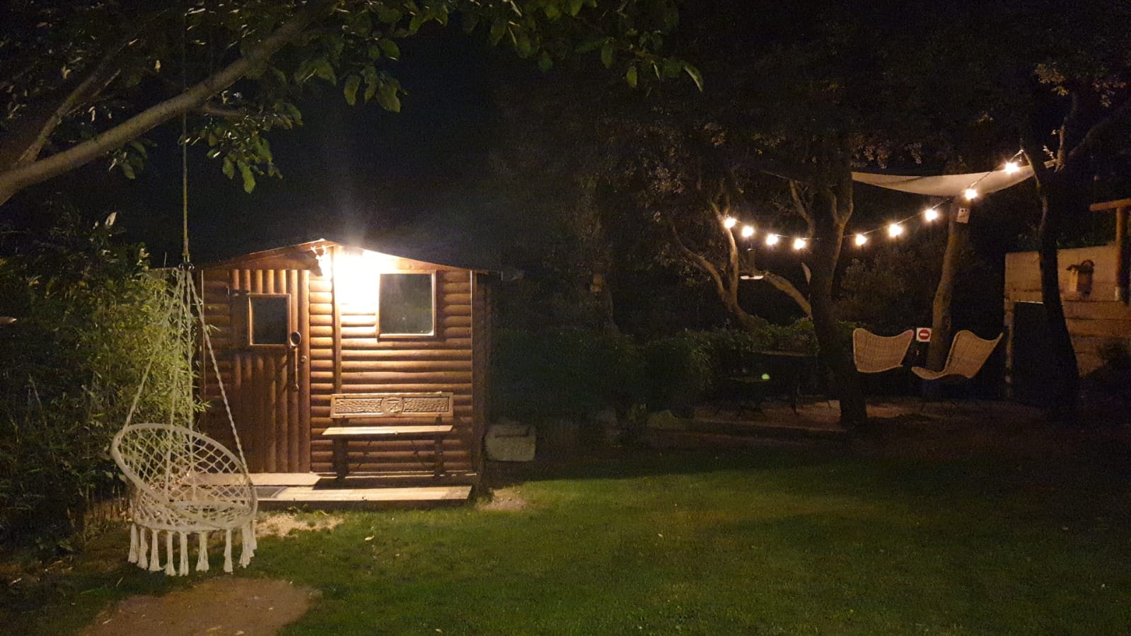 Escena nocturna d'un pati del darrere amb una caseta de fusta i llums de corda que il·luminen un gronxador . Cal Serni