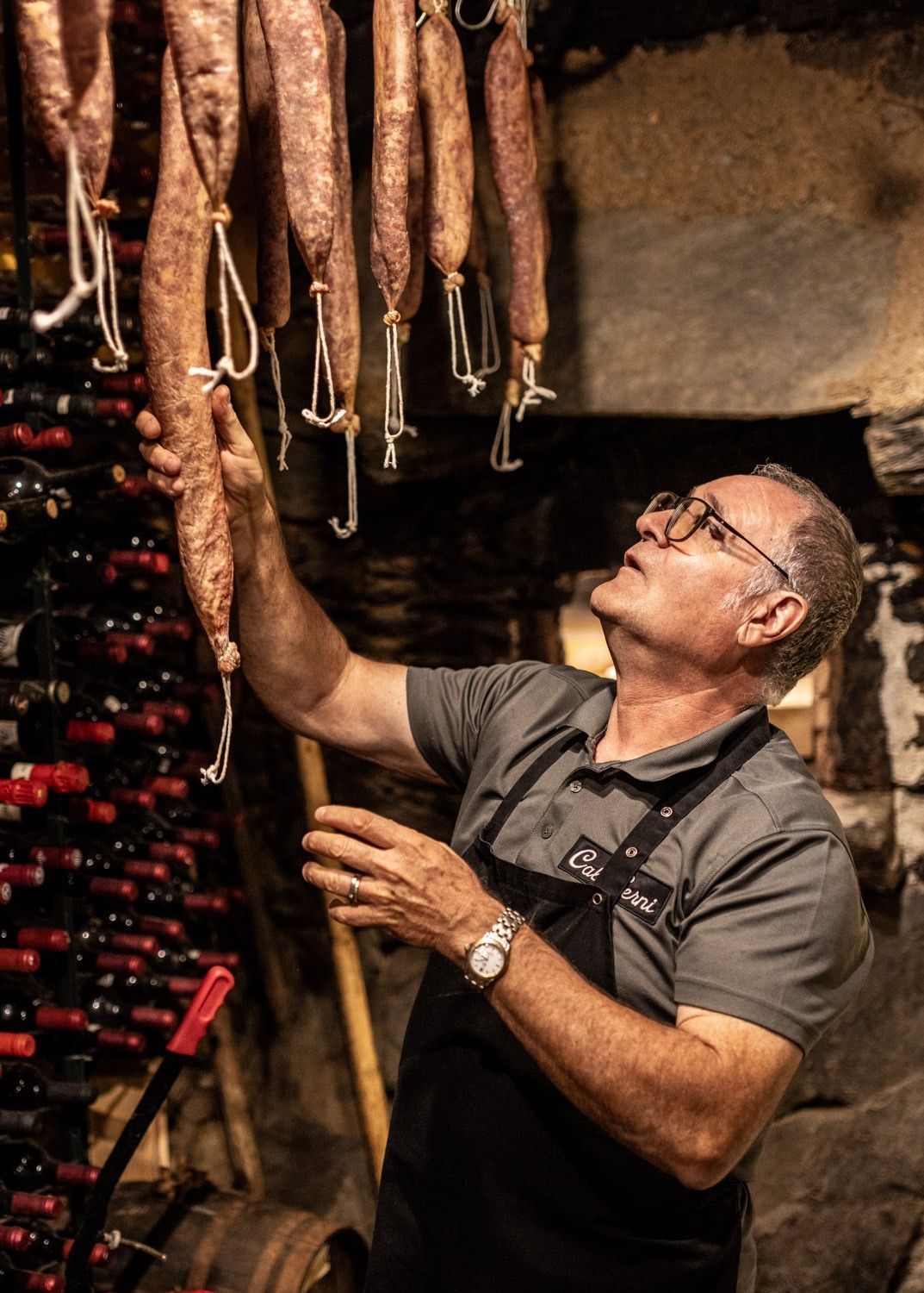 Home examinant salsitxes curades penjades en un celler ple d'ampolles de vi. Cal Serni