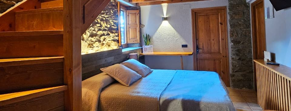 Dormitori acollidor amb un llit, escala i detalls de paret de pedra. Cal Serni