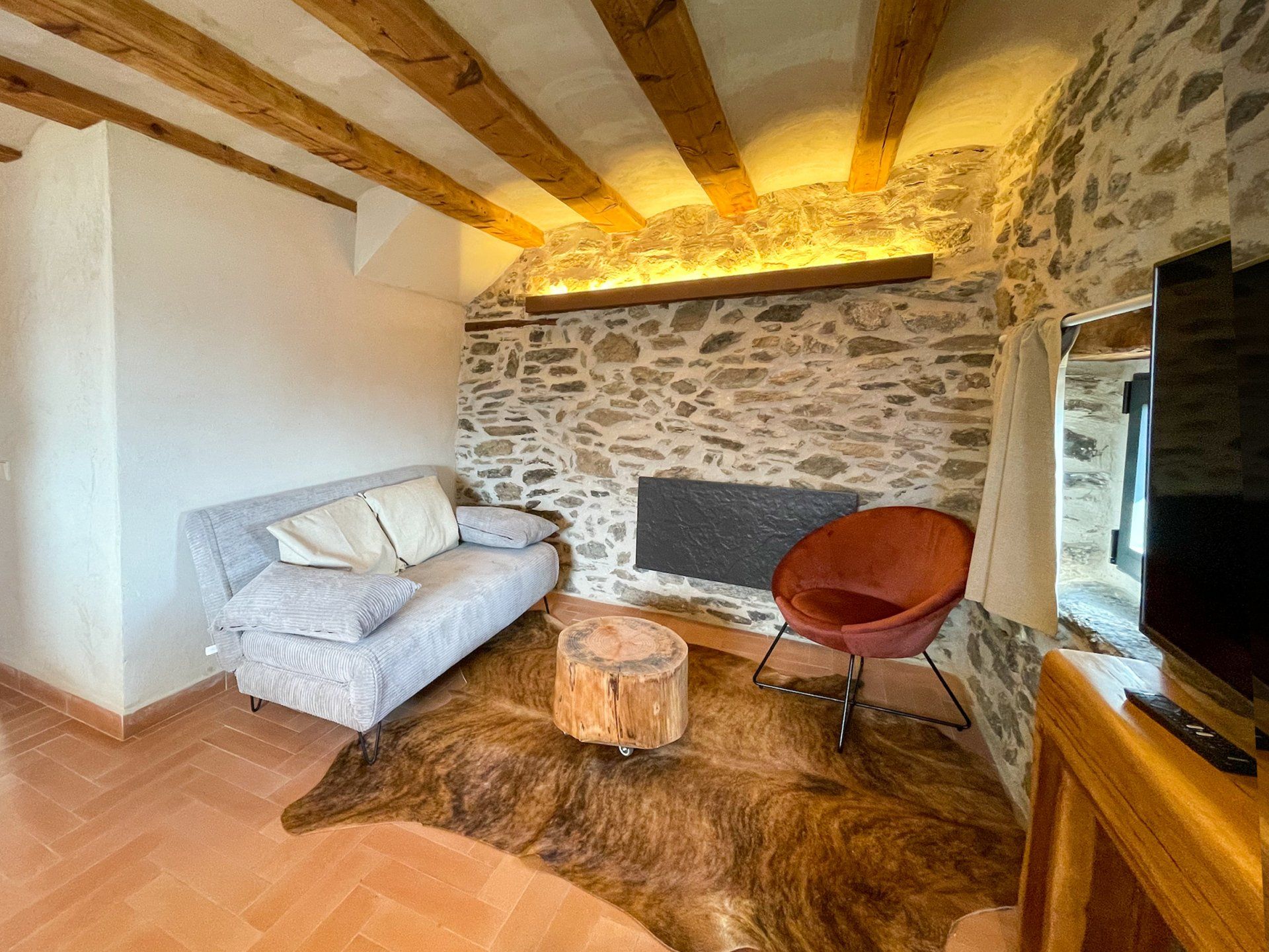 Sala d'estar amb paret de pedra, bigues de fusta, sofà, cadira i catifa. Cal Serni