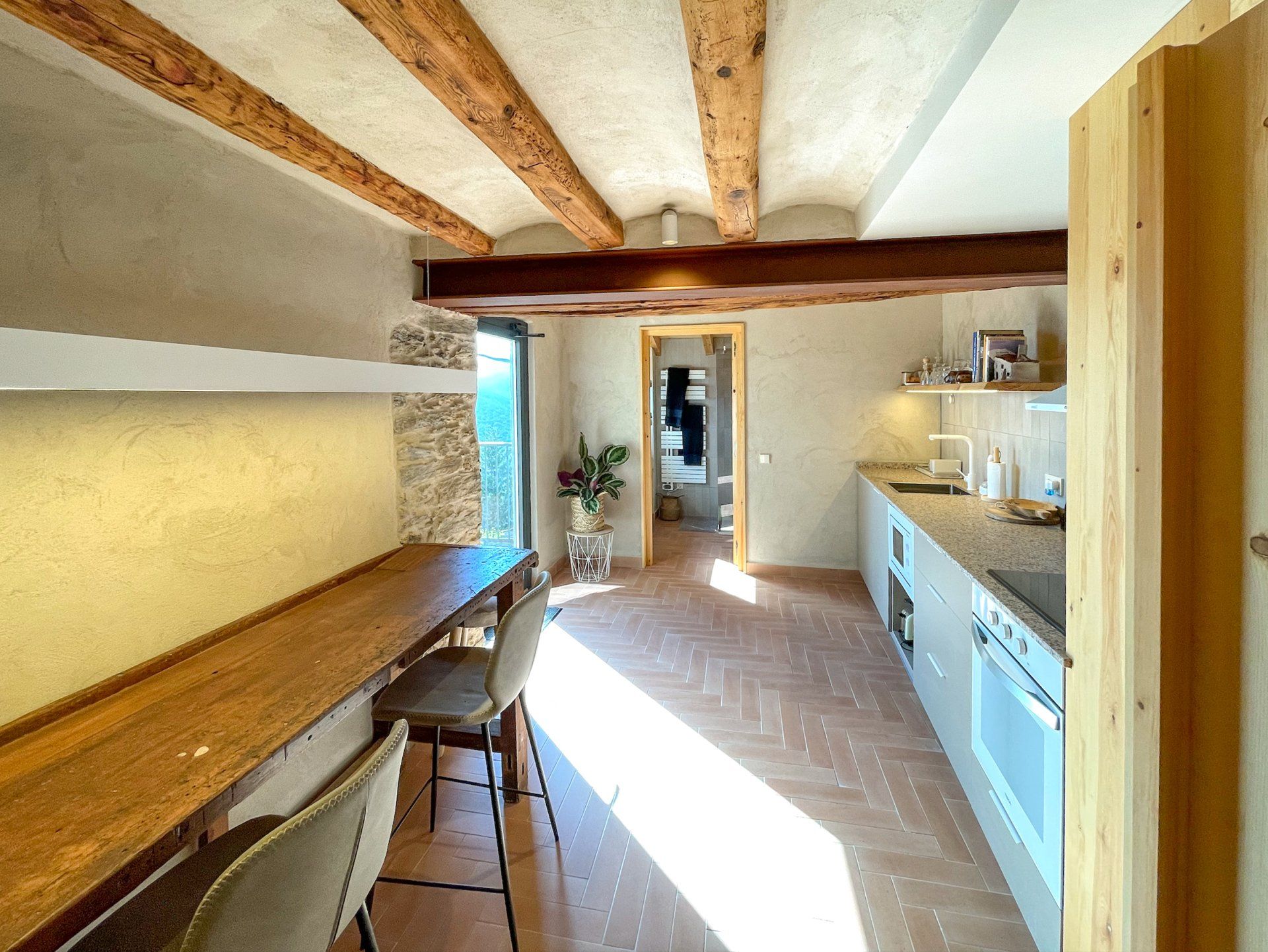 Cuina interior amb bigues de fusta, taula llarga de fusta amb dues cadires i armaris de colors clars. Cal Serni