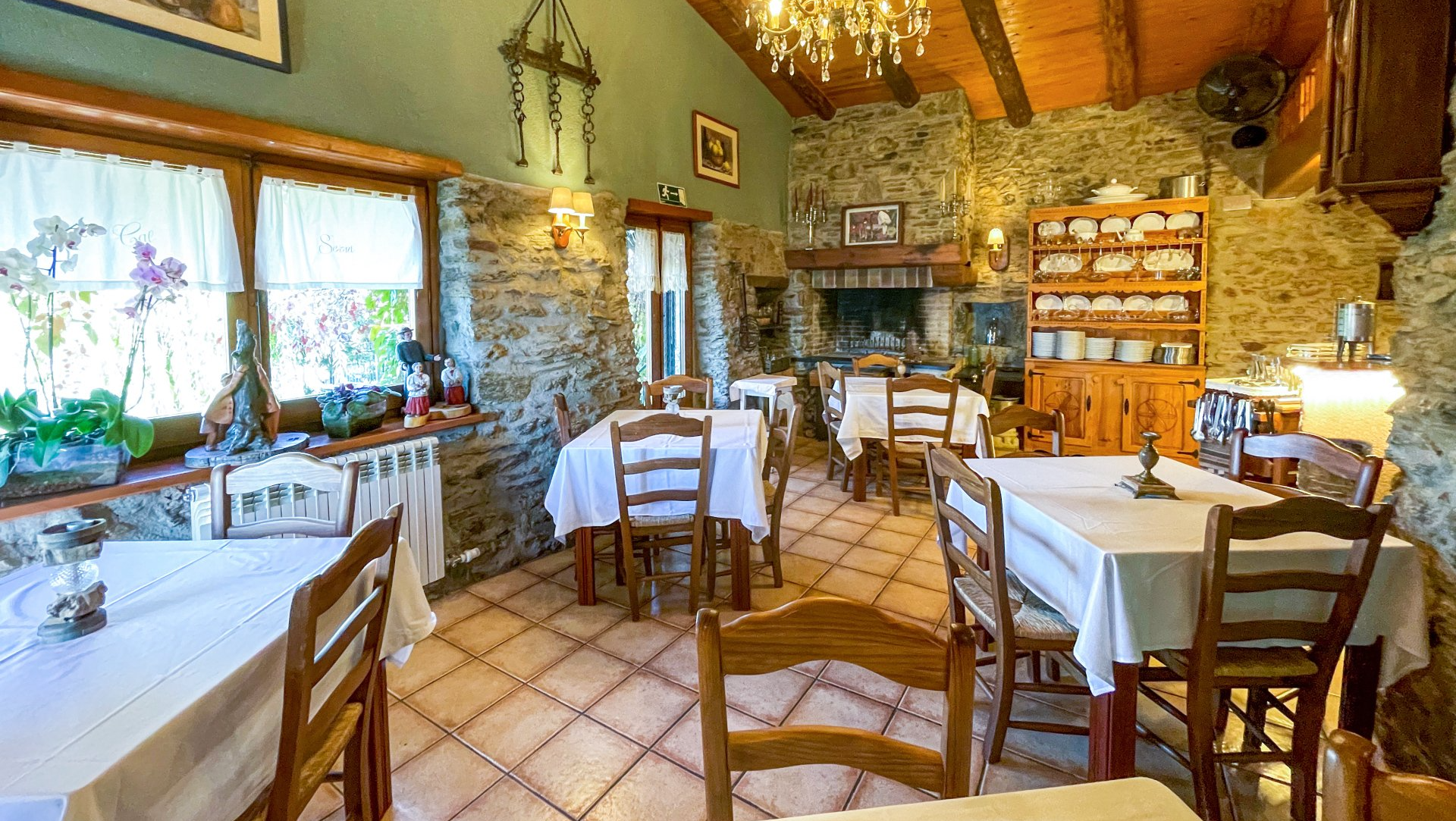 Interior de restaurant amb parets de pedra, cadires de fusta, estovalles blanques i llar de foc. Cal Serni