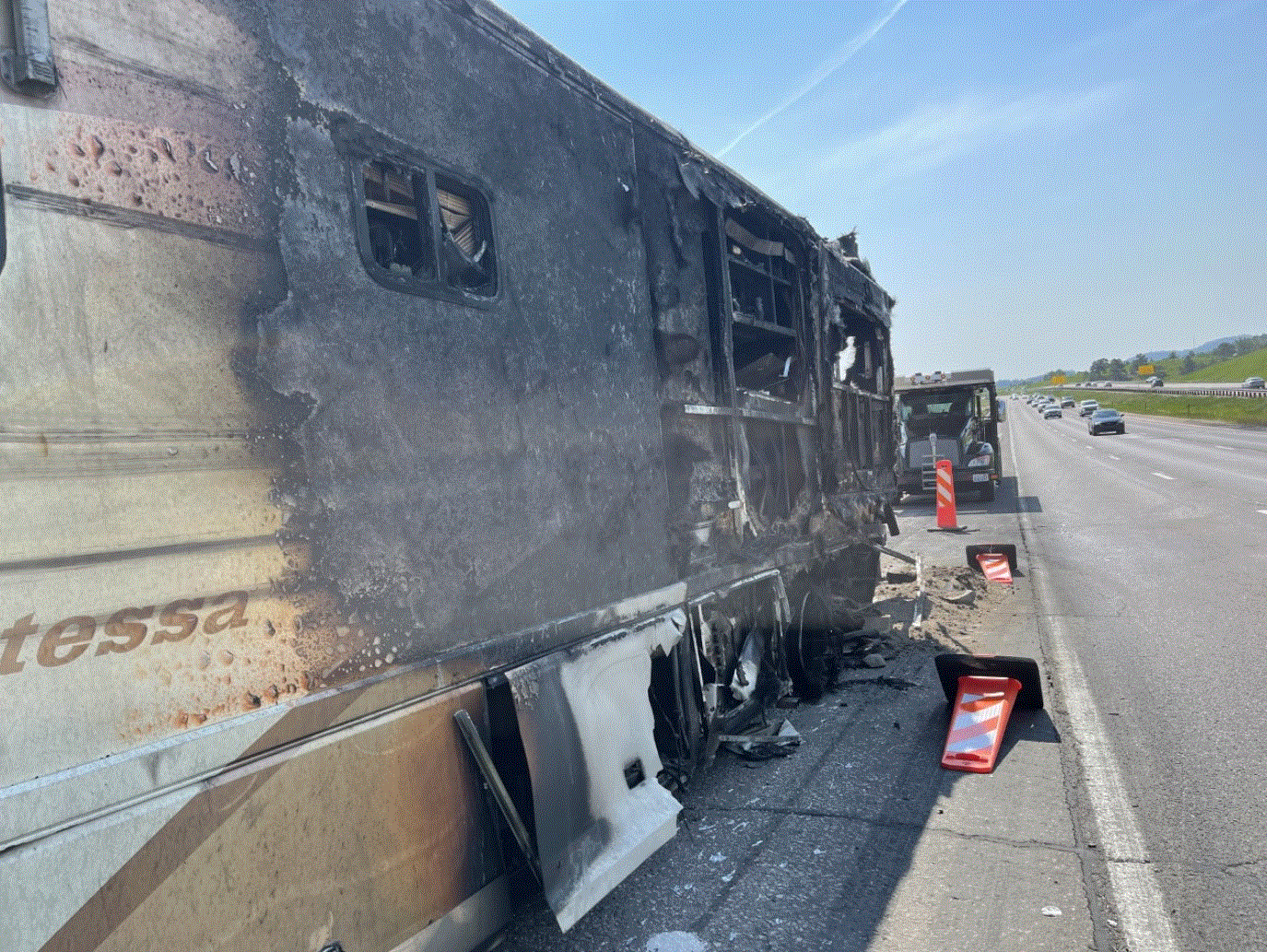 Burnt RV parked on the shoulder of a highway, damage from fire visible.