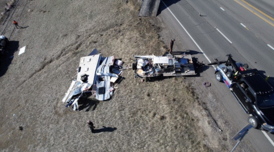 Overturned RV wreckage beside a highway, with tow truck and people nearby.