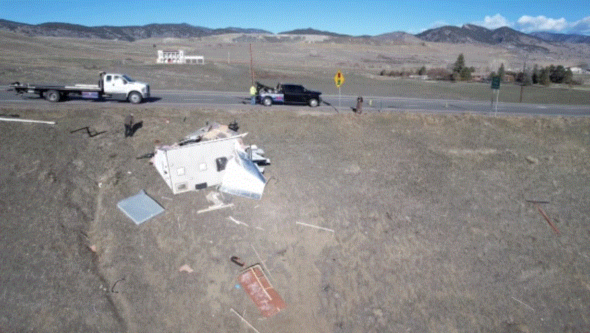 Wrecked white vehicle off road; two tow trucks present. Mountains in background, sunny day.