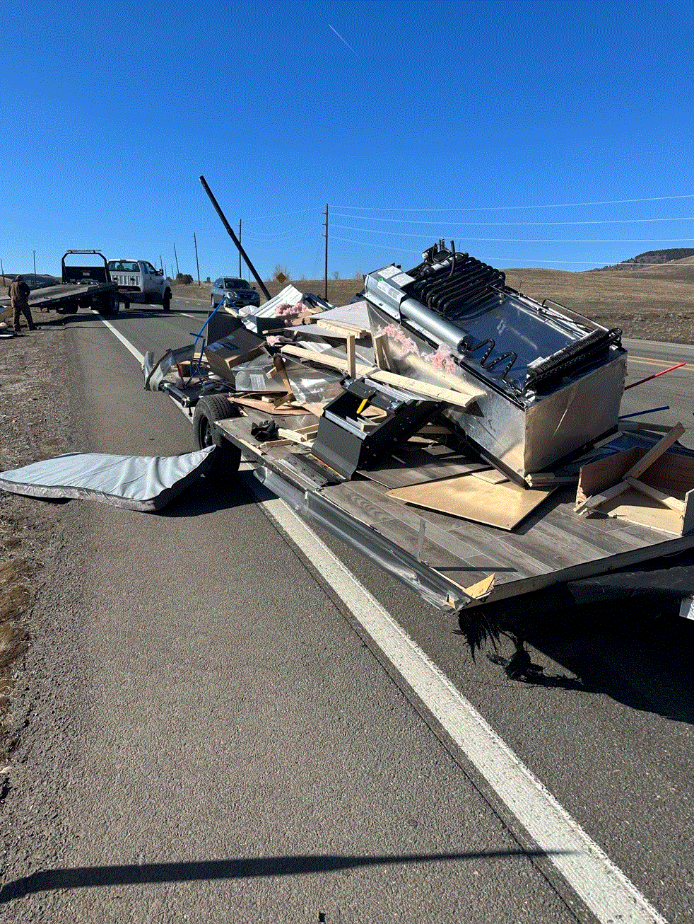 Overturned trailer on a road with debris, containing metal and wooden objects. A tow truck is present.