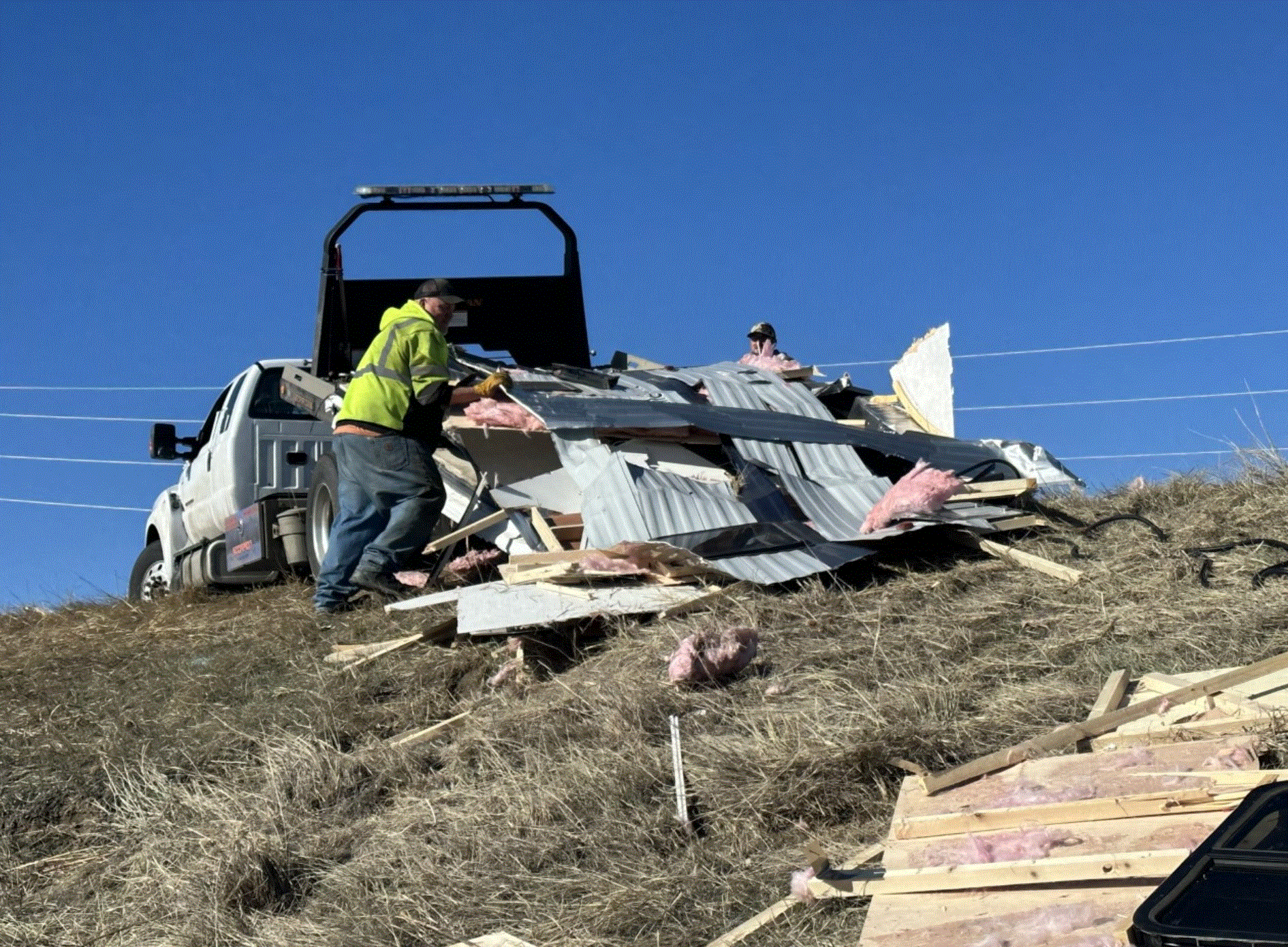 Man in a high-vis vest loads debris from a collapsed structure onto a truck, on a hillside.