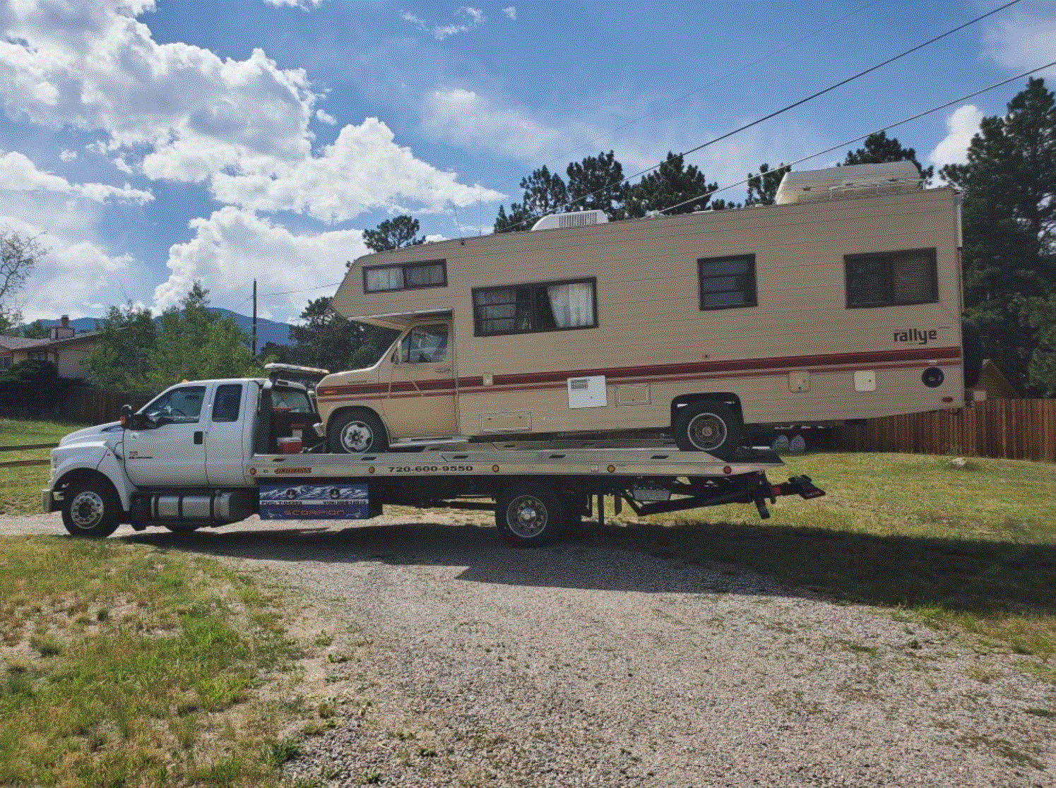 A beige RV being towed by a white tow truck on a gravel road, under a cloudy sky.