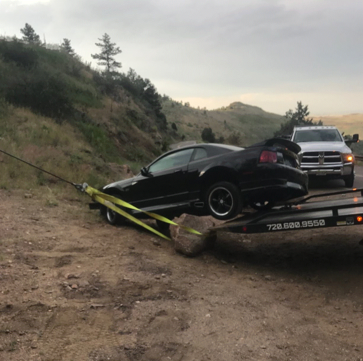 Black car being towed onto a flatbed by a white tow truck on a dirt road next to a hill.