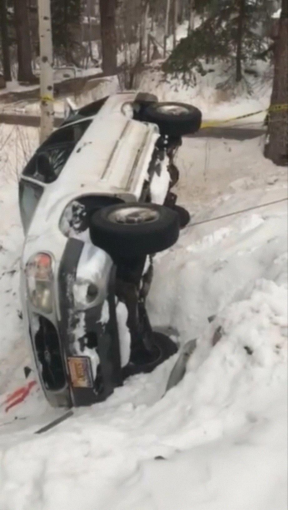 White car overturned in snow-filled ditch, headlights visible, in a wooded area.