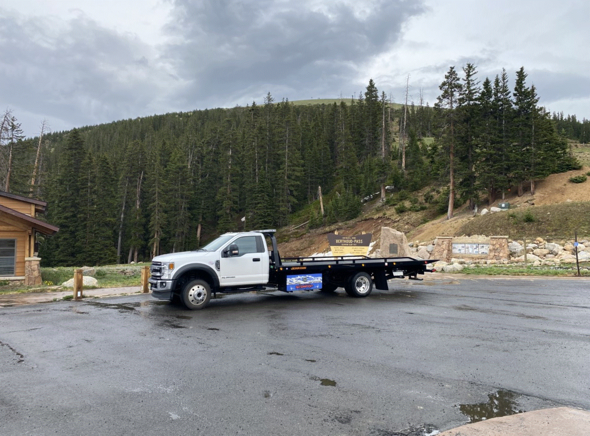 White flatbed truck parked in a wet parking lot, with a forested mountain in the background under a cloudy sky.