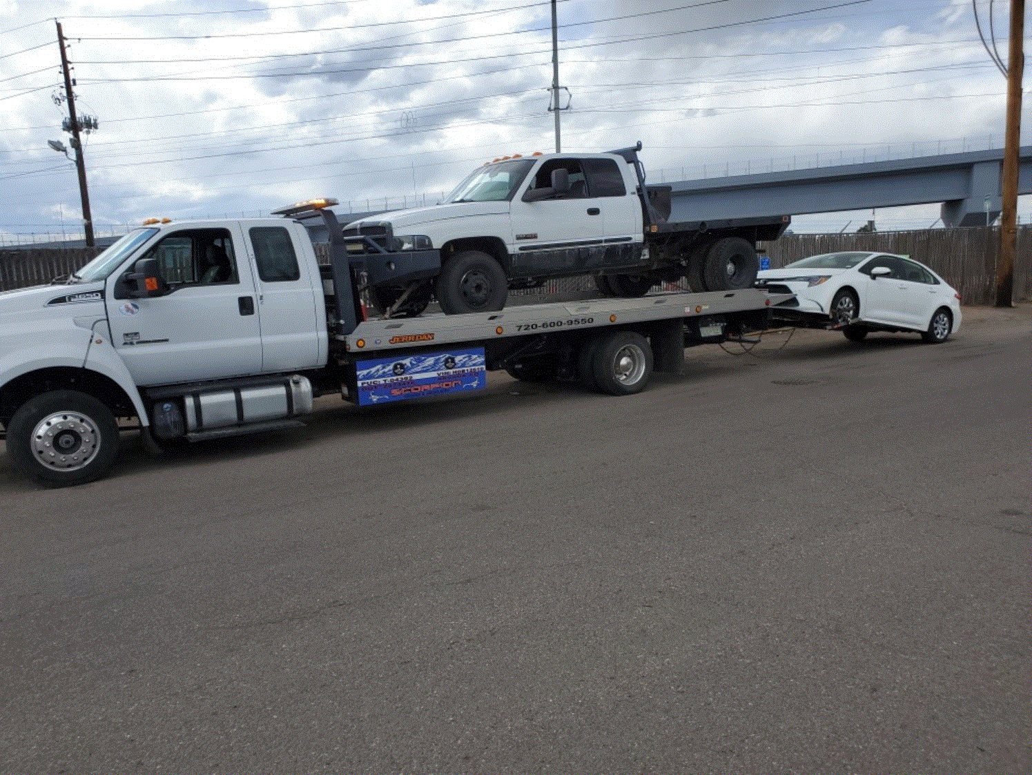 White tow truck hauling a white pickup truck and a white car on a city street under a cloudy sky.