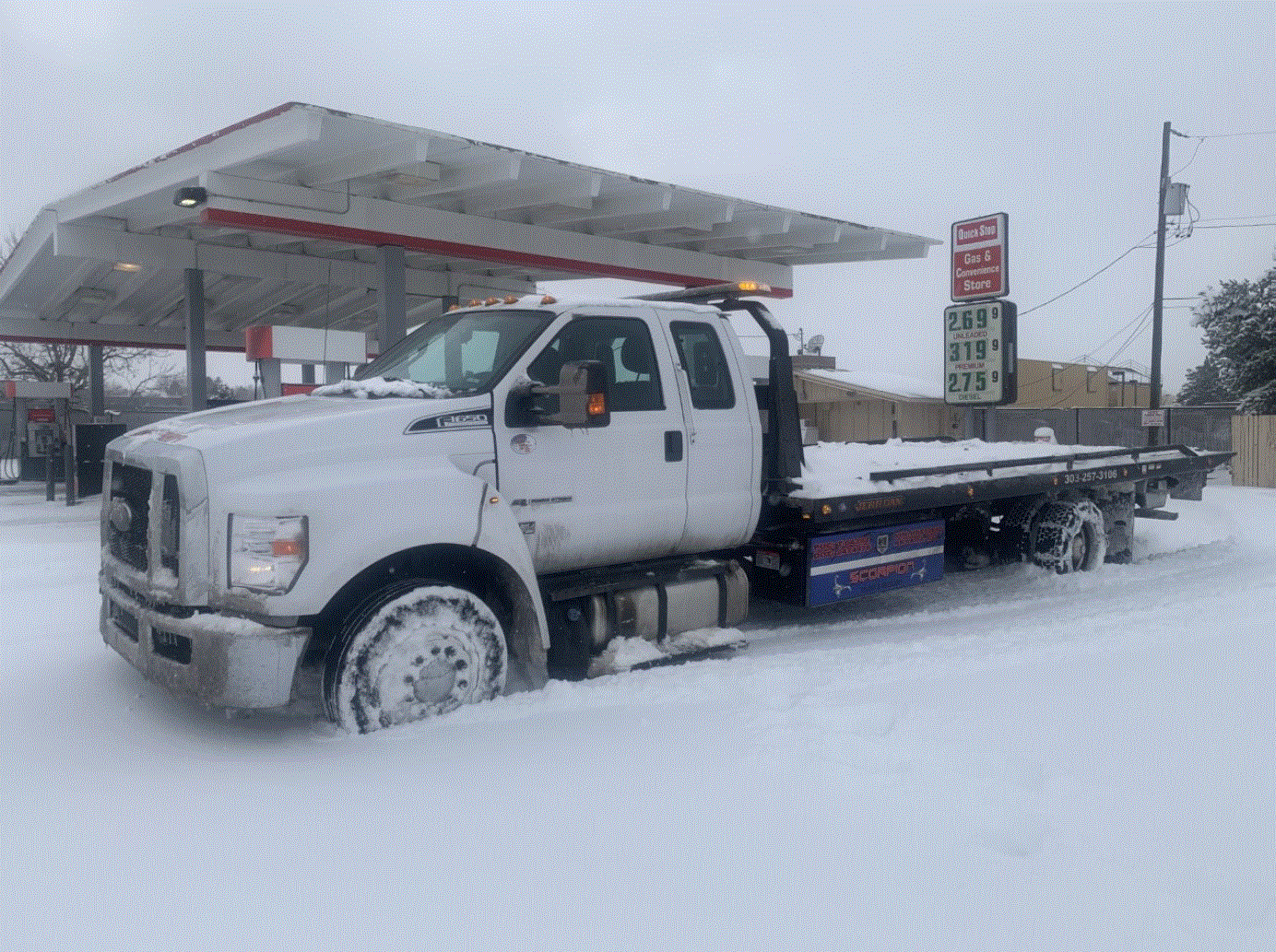 White tow truck stuck in snow at a gas station with prices on a sign.