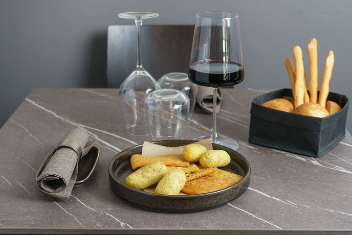 Tavola apparecchiata con grissini, pasticcini, vino, bicchieri capovolti e un tovagliolo arrotolato su un tavolo di marmo grigio.