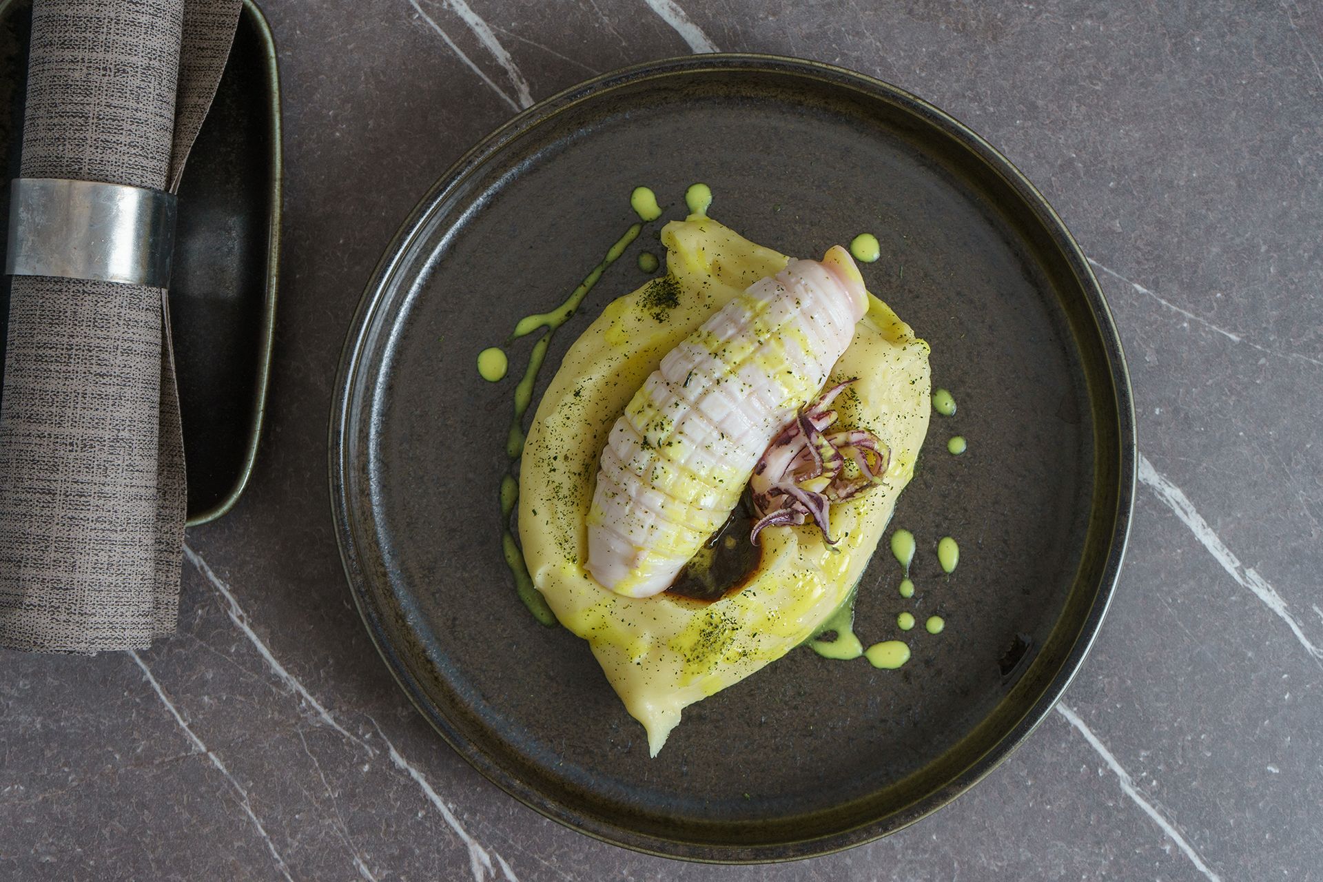 Filetto di pesce servito su salsa verde, con coltello e forchetta su un tavolo grigio.