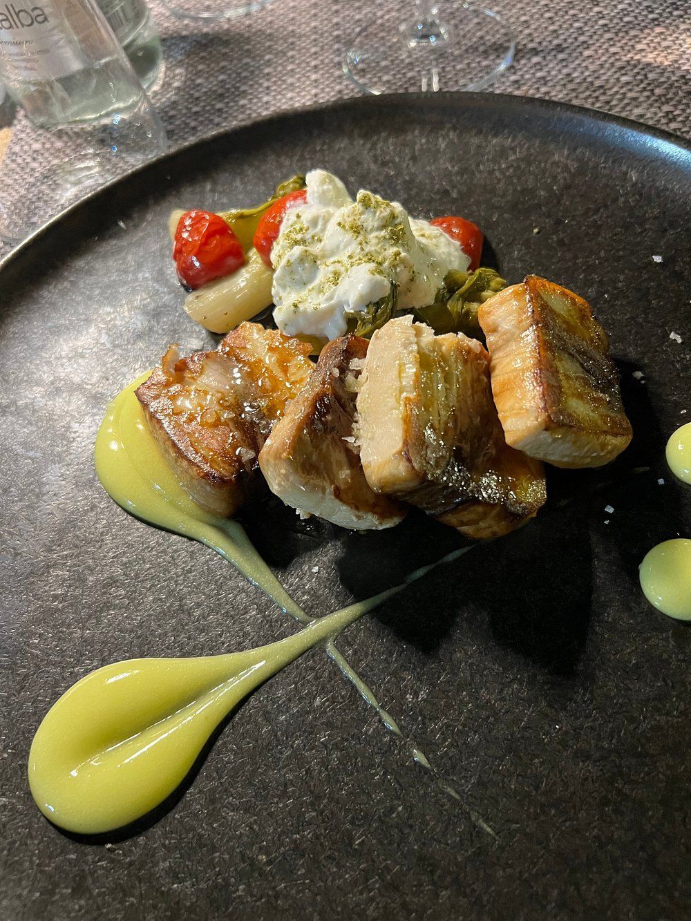Pesce scottato servito su un piatto scuro con salsa cremosa, pomodori e guarnizione di erbe aromatiche.