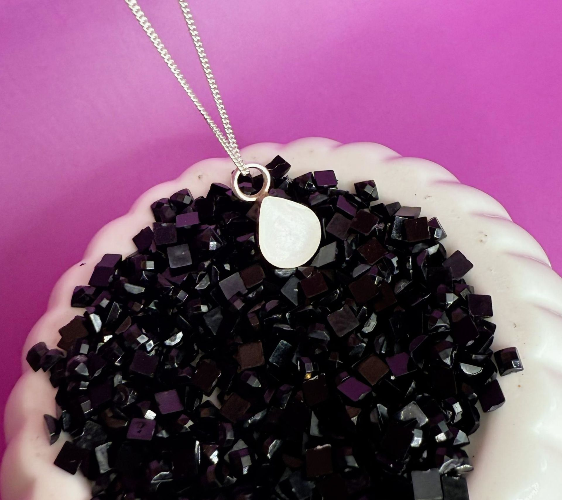 Silver necklace with a white pendant resting on a bowl of black beads against a pink background