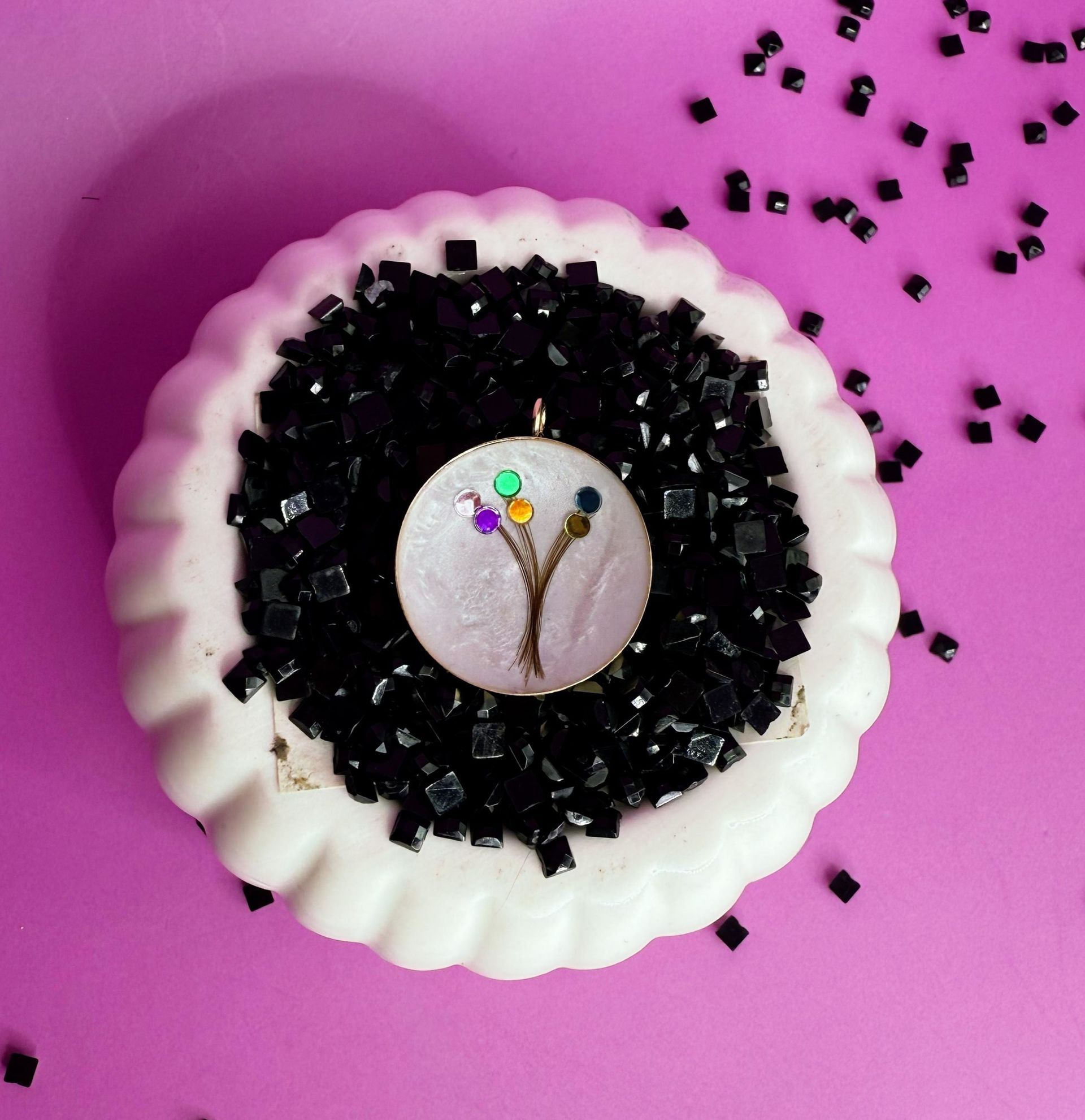 White scalloped dish with black crystals and a small jeweled ornament on a pink background