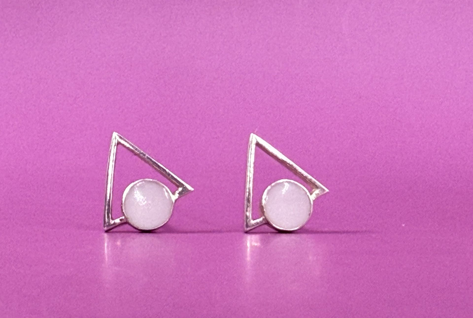 Two silver geometric earrings with white beads on a pink background
