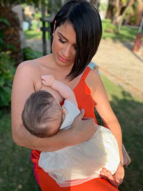 Woman holding a baby outdoors on a sunlit path, looking down with a gentle expression.
