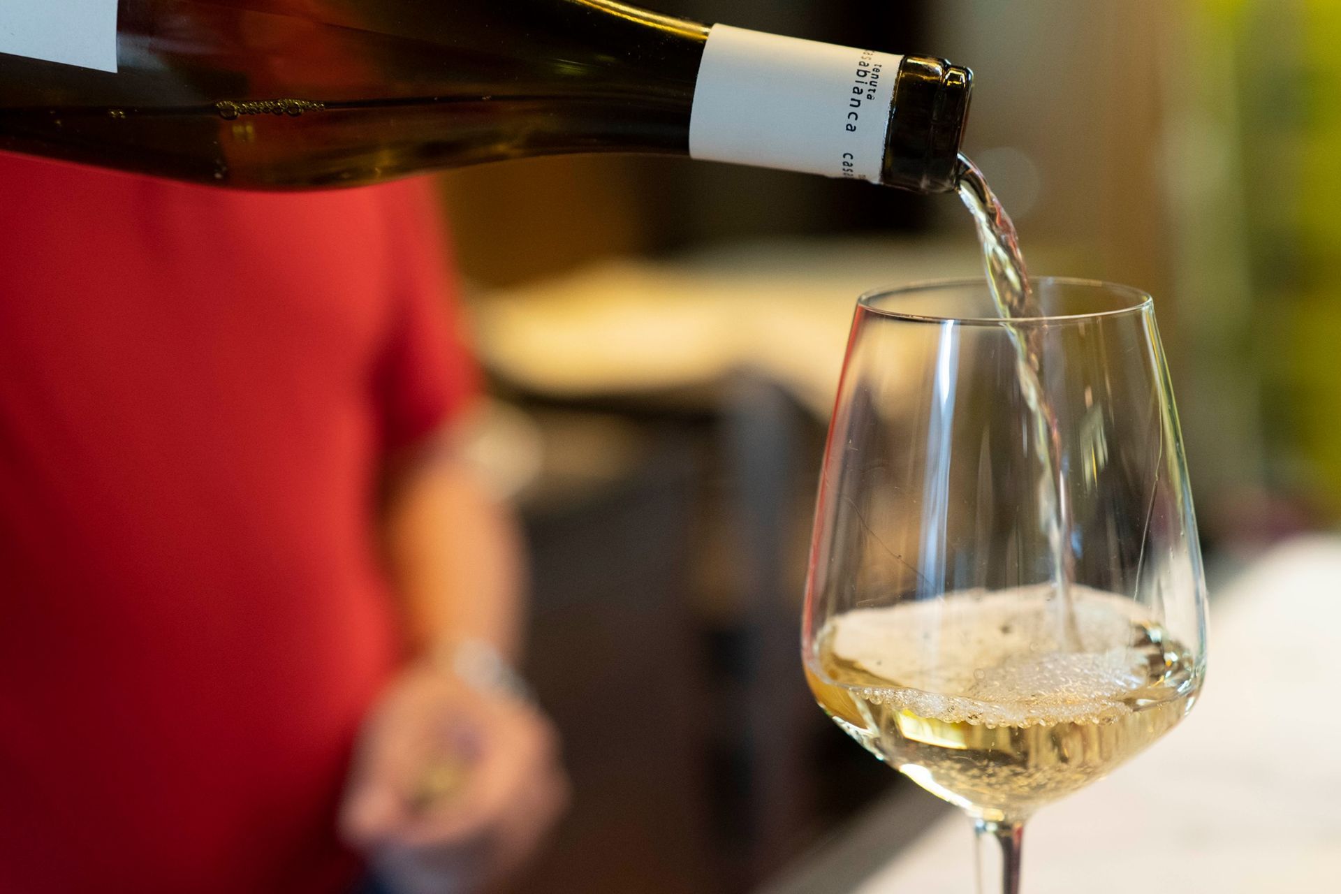 Vino versato da una bottiglia in un bicchiere; sullo sfondo una camicia rossa.