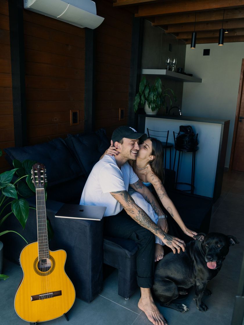 Duas pessoas sentadas em um sofá com um cachorro preto em uma sala rústica, com um violão encostado por perto.